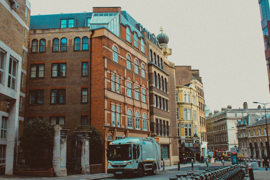 A medium-sized commercial refuse collection vehicle is parked on the street in front of a historic, multi-storey brick building with large, arched windows and detailed stonework in the heart of Knight