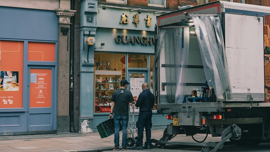 Two movers from Knightsbridge Man and Van are preparing a home relocation outside a shop with Chinese signage, with one individual holding a green plastic crate and the other guiding a dolly. The loading process involves a large, open cardboard box on a trolley positioned on the pavement, close to a moving truck with its side curtain partially pulled back to reveal the interior. The scene takes place in daylight, with the storefront behind them displaying various items inside, and the exterior features typical urban architecture with decorative molding around windows and doors. The movers are dressed in dark clothing and are engaged in organized packing and furniture transport activities, illustrating the meticulous steps involved in residential moving and packing services in Knightsbridge.