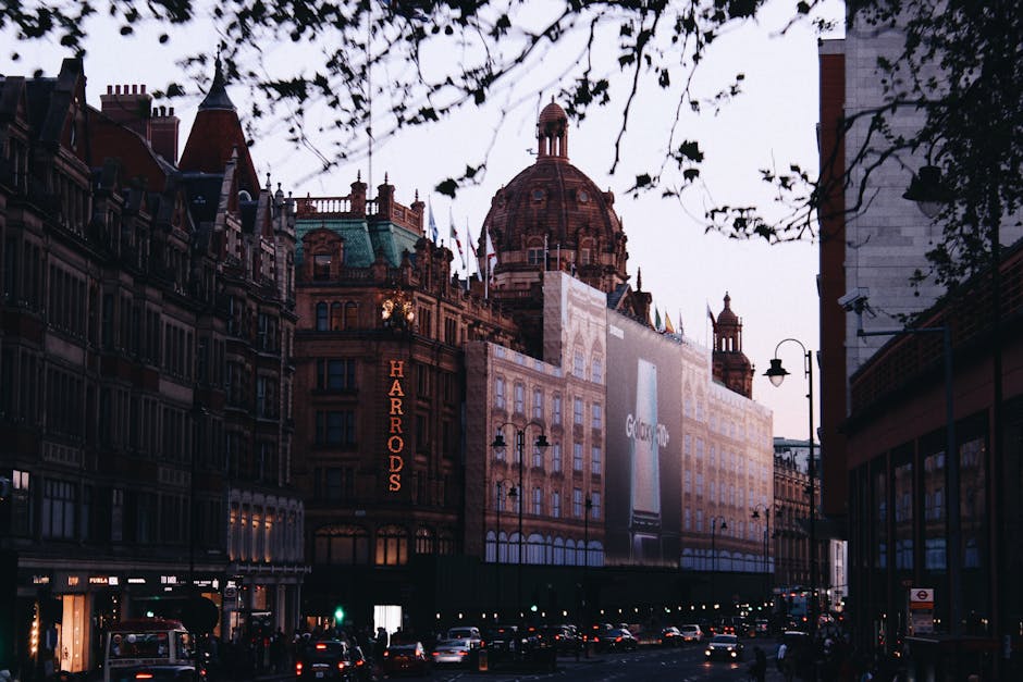 A street view in Knightsbridge with the Harrods department store prominently visible, featuring its illuminated vertical sign and ornate architecture topped by a large, domed roof. Surrounding buildings include multi-story structures with classic brick facades and decorative elements, some partially covered by white scaffolding and protective sheeting for ongoing refurbishment or delivery access preparations. The scene shows cars parked along the pavement and a few moving vehicles, with street lamps and a billboard visible on the building façade. The image is captured during dusk or early evening, with a dim sky and a slightly overcast atmosphere. This setting highlights the urban environment where professional removals services, such as those provided by Knightsbridge Man and Van, are often involved in logistical tasks like home relocation and furniture transport, especially in such busy shopping and residential districts.