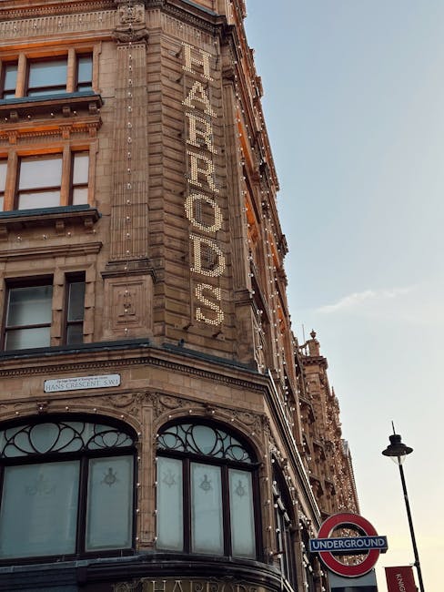 Close-up view of the exterior corner of a historic multi-storey building in the Harrods area of Knightsbridge, featuring intricate architectural details and large windows on the lower floors. The building's facade displays a vertical sign spelling 'FASHION' in illuminated letters, attached to the stone wall. The sky is clear with gentle daylight illuminating the structure. A traditional street lamp and an underground public transport sign are visible in the foreground, indicating a busy urban environment. The scene captures the classic architectural style typical of central London, relevant to house removals and furniture transport services provided by Knightsbridge Man and Van for home relocation and moving logistics.