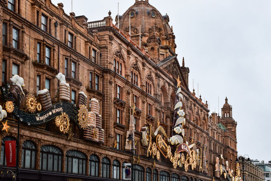 A street view in Knightsbridge with the Harrods department store prominently visible, featuring its illuminated vertical sign and ornate architecture topped by a large, domed roof. Surrounding buildings include multi-story structures with classic brick facades and decorative elements, some partially covered by white scaffolding and protective sheeting for ongoing refurbishment or delivery access preparations. The scene shows cars parked along the pavement and a few moving vehicles, with street lamps and a billboard visible on the building façade. The image is captured during dusk or early evening, with a dim sky and a slightly overcast atmosphere. This setting highlights the urban environment where professional removals services, such as those provided by Knightsbridge Man and Van, are often involved in logistical tasks like home relocation and furniture transport, especially in such busy shopping and residential districts.