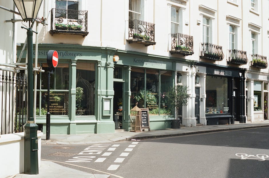 A street scene in Kensington and Chelsea featuring a row of commercial premises, including a florist shop with a pale green facade and large display windows filled with plants and flowers. The shop has decorative architectural details and a small blackboard sign placed outside on the sidewalk. Adjacent to the florist is a gallery with a black exterior. The sidewalk is paved with concrete, and a white striped crosswalk marking is visible on the road, which appears to be a quiet urban street. A streetlamp and metal posts are positioned near the shop entrance. The buildings above the shops are white with multiple floors, each with black wrought iron balconies decorated with potted plants. The lighting indicates a clear day, and the scene provides a neutral, factual view of a typical London street suitable for home relocation or furniture transport processes, often managed by companies like Knightsbridge Man and Van.