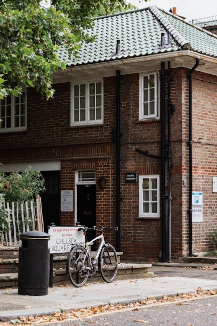 Outdoor view of a brick residential building with a tiled, gabled roof and white-framed windows. In front of the building, a paved sidewalk features a black trash bin, a bicycle secured to a metal bike rack, and a sign indicating residential parking restrictions in the area. Partially visible greenery from trees provides shade over the scene. The image captures a typical urban street scene relevant to home relocation and moving logistics, with the building’s entrance and accessible sidewalk for loading and temporary storage during furniture transport, as handled by Knightsbridge Man and Van.