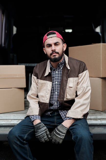 A young man with a beard, wearing a red cap, a brown and beige jacket over a checkered shirt, and striped gloves, is sitting at the back of a van with the tailgate lowered. Behind him, there are several stacked cardboard boxes, some wrapped in plastic or tissue paper, indicating packing materials used during a house removal. The interior of the van appears spacious and is used for transport of household items. The van is parked outside a property, with a dark environment suggesting early morning or late evening lighting. The scene illustrates the loading process involved in home relocation, supported by a professional removals service like Knightsbridge Man and Van. The image captures the moment before or after furniture and boxes are moved, highlighting aspects of furniture transport and packing during the moving process in London’s SW1X area.