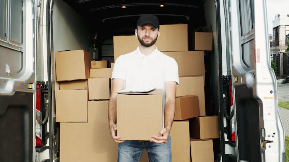 A man wearing a white polo shirt and a black cap stands inside the open cargo area of a white van, holding a cardboard box with both hands. Behind him, numerous additional cardboard boxes of varying sizes are stacked neatly within the van, some wrapped in plastic or placed directly on the floor. The van is parked outside a residential building, with part of a sidewalk and garden visible on the right side of the image. The scene is well-lit, indicating daytime, and the setting suggests an active home relocation or furniture transport process as part of a professional removal service by Knightsbridge Man and Van, specialising in house removals and moving logistics for SW1X flats.