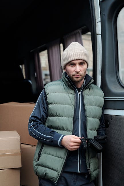 A young man with a beard, wearing a red cap, a brown and beige jacket over a checkered shirt, and striped gloves, is sitting at the back of a van with the tailgate lowered. Behind him, there are several stacked cardboard boxes, some wrapped in plastic or tissue paper, indicating packing materials used during a house removal. The interior of the van appears spacious and is used for transport of household items. The van is parked outside a property, with a dark environment suggesting early morning or late evening lighting. The scene illustrates the loading process involved in home relocation, supported by a professional removals service like Knightsbridge Man and Van. The image captures the moment before or after furniture and boxes are moved, highlighting aspects of furniture transport and packing during the moving process in London’s SW1X area.