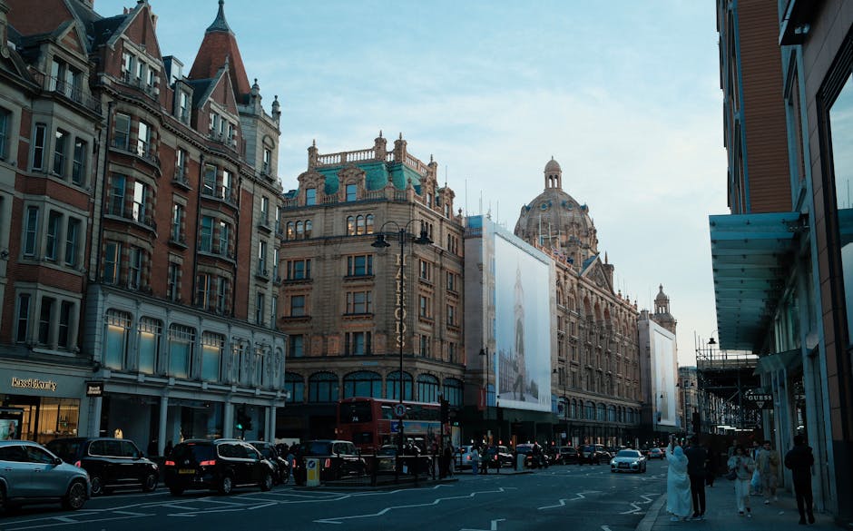 A city street scene outside a historic building with ornate architectural details and a prominent dome, captured during daytime with natural lighting. The street is busy with parked cars along the curb, pedestrians walking on the sidewalk, some wearing formal attire, and a double-decker bus visible in the background. On the right side, a modern glass storefront can be seen, with scaffolding and construction materials suggesting ongoing work. The sky is clear with a few clouds. This setting illustrates an urban environment suitable for house removals or furniture transport, as handled by Knightsbridge Man and Van, including loading or moving activities occurring in proximity to the buildings and street environment.