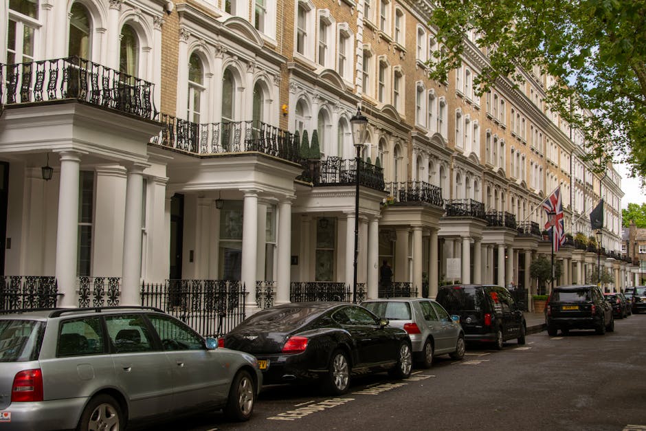 A row of elegant Victorian-style terraced houses with white facades, decorative columns supporting balconies, black wrought iron railings, and large windows with intricate trim, located on a busy street with parked cars including a silver estate, a black coupe, and several other vehicles. The street features black lamp posts, flags including a Union Jack, and lush green trees providing shade. The scene is captured in daylight, with the façade of residential properties visible behind the parked vehicles, suitable for a professional home relocation or furniture transport service such as Knightsbridge Man and Van, as part of packing and moving processes.
