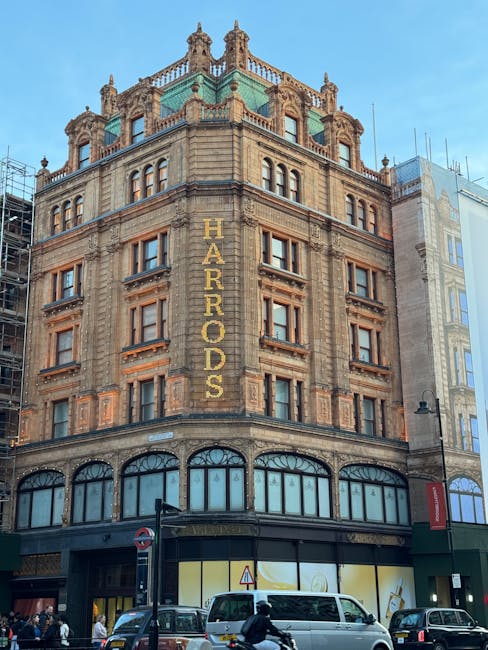 A city street scene outside a historic building with ornate architectural details and a prominent dome, captured during daytime with natural lighting. The street is busy with parked cars along the curb, pedestrians walking on the sidewalk, some wearing formal attire, and a double-decker bus visible in the background. On the right side, a modern glass storefront can be seen, with scaffolding and construction materials suggesting ongoing work. The sky is clear with a few clouds. This setting illustrates an urban environment suitable for house removals or furniture transport, as handled by Knightsbridge Man and Van, including loading or moving activities occurring in proximity to the buildings and street environment.