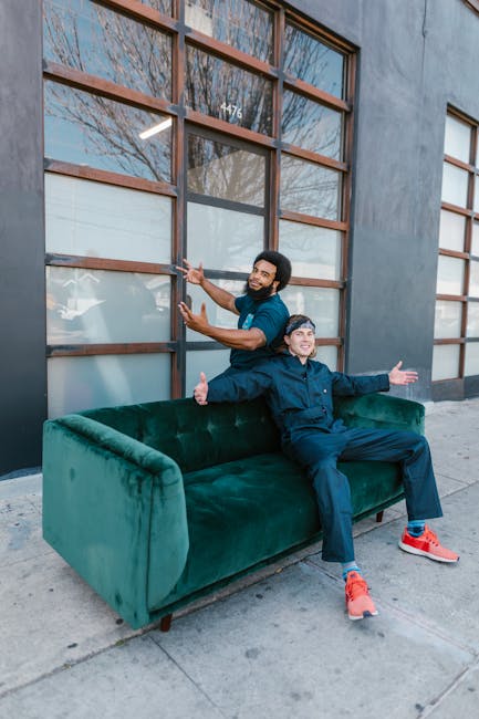 Two men are positioned outside a building with large wooden-framed windows, engaging in a casual, cheerful moment during a home relocation process. One man, standing behind a green velvet sofa placed on the sidewalk, is smiling and gesturing with his hands, possibly indicating the transport or movement of furniture. The other man is seated on the sofa, facing forward with both arms extended outward in a welcoming gesture, and is dressed in dark blue clothing with bright orange sneakers. The setting appears to be in an urban area, with a concrete pavement and a building featuring multiple rectangular, wooden window panels behind them. The scene suggests an informal moment during furniture transport or packing and moving preparations, with the sofa likely being part of a larger collection of belongings for a house removal, supported by a professional moving company such as Knightsbridge Man and Van.